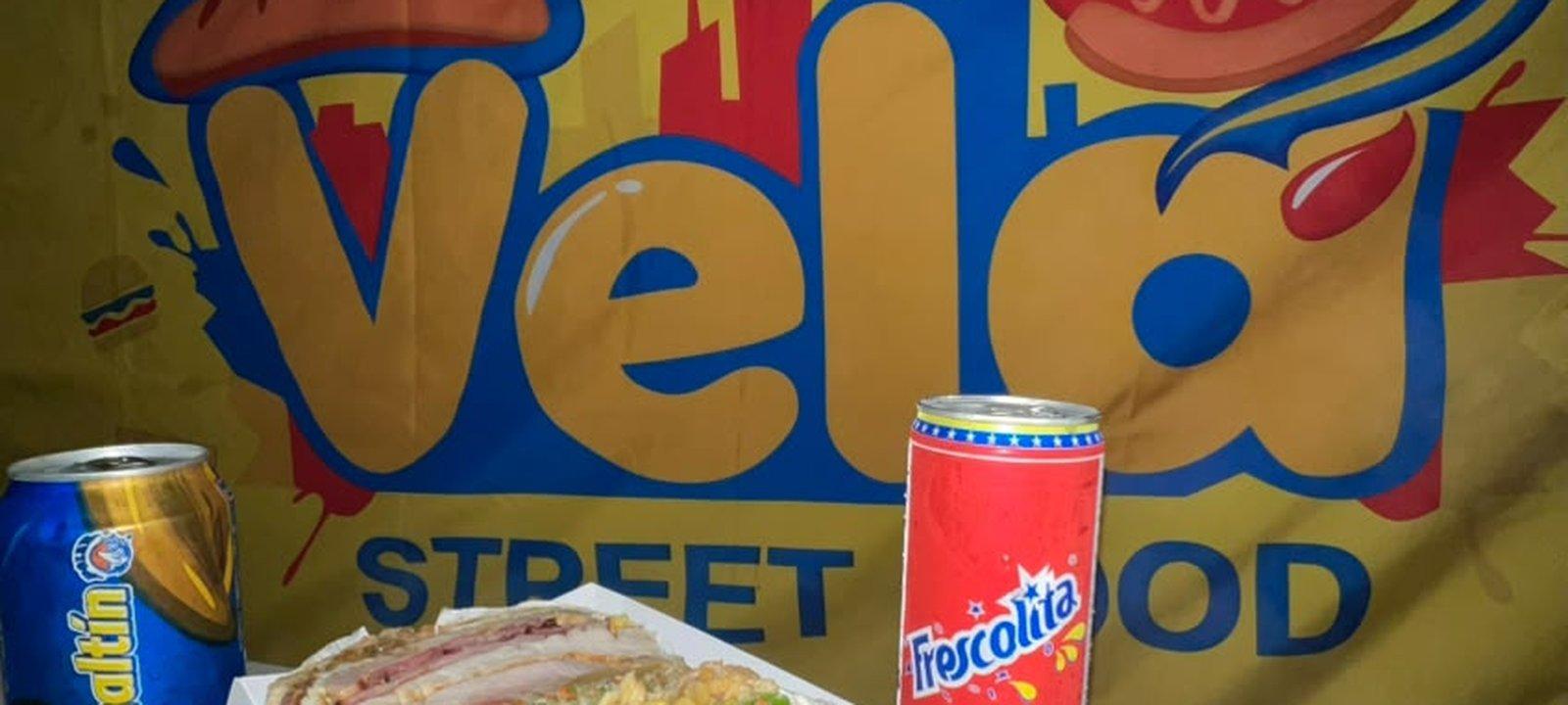 A Hamburguesa Triple with a Malta and a Frescolita in front of the Vela Street Food banner