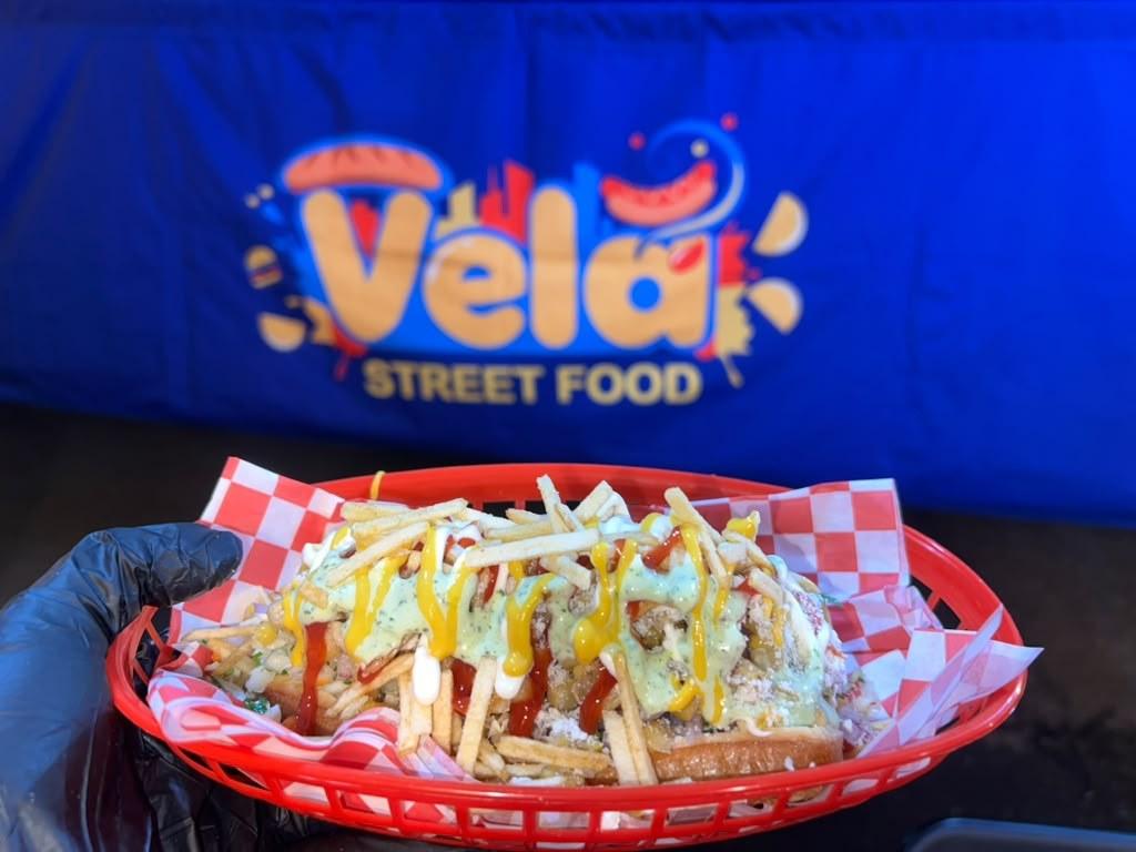 A Perro Normal in a red basket in front of the Vela Street Food banner, covered in potato sticks and sauces